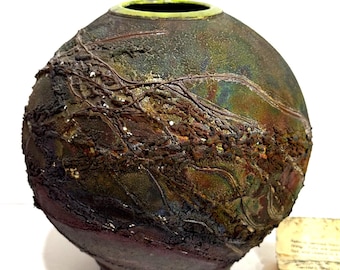 May include: A handcrafted ceramic vase with a textured, organic design. The vase features a spherical shape with a dark, earthy color palette of browns, greens, and purples. The rim is accented with a bright green color. The vase sits on a dark base.