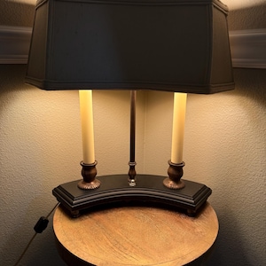 May include: A dual-light table lamp featuring a rectangular black fabric shade and two cream-colored candle-style bulbs. The base is a curved black platform with two bronze-toned candle holders and a central metal rod, emitting a warm glow.