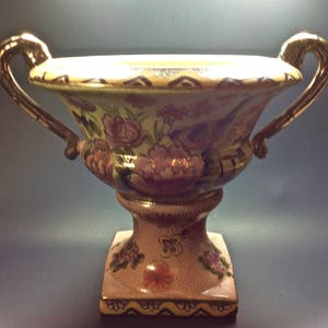 May include: A ceramic urn-style vase featuring two gold-toned handles, a wide flared rim, and a square base. The body is decorated with painted pink and purple floral motifs against a cream background, with additional floral patterns on the pedestal.
