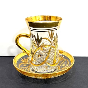 May include: A clear glass teacup and saucer set with gold trim and accents. The teacup has a handle and is decorated with gold leaf patterns. The saucer mirrors the teacup's design, creating a cohesive and elegant look. The set is designed for serving tea.