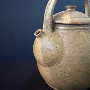 May include: A handmade, beige ceramic teapot with a curved handle and a lid. The teapot has a rounded body and a spout. The handle and lid are the same color as the body. The teapot is likely used for brewing tea.