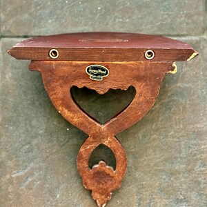 Puede incluir: Una estanter&iacute;a de pared de madera vintage con un dise&ntilde;o recortado en forma de coraz&oacute;n. La estanter&iacute;a es de color marr&oacute;n c&aacute;lido con un soporte inferior decorativo. La marca "Syroco Wood" es visible en una peque&ntilde;a etiqueta.