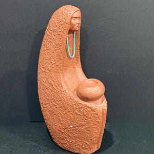 May include: A terracotta-colored clay sculpture depicting a figure wrapped in a textured cloak, holding a small rounded vessel in their lap. The figure wears a bright turquoise beaded necklace around their neck, contrasting with the earthy, rough-surfaced clay material of the statue.