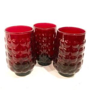 May include: Three cylindrical drinking glasses made of deep red glass. Each glass features a repeating pattern of raised, circular bubble-like indentations covering the exterior surface, with a smooth rim and a solid, flat base for stability.