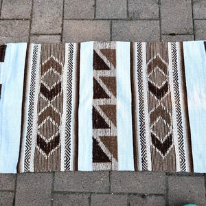 May include: A rectangular woven rug with a geometric pattern in shades of brown, white, and cream. The rug has a central section with brown triangles and vertical stripes. The edges are finished with tassels.