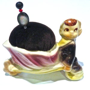 May include: A vintage ceramic pin cushion in the shape of a boy pushing a shell-shaped cart. The cart holds a round, black velvet cushion. A decorative pin with a black bead, red beads, and a pearlescent oval tops the cushion. The figurine is painted in shades of brown, yellow, and pink.