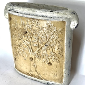 May include: A rectangular ceramic vase with a distressed white finish on the edges and a central tan panel featuring a raised relief design of a tree with leaves, flowers, and small fruits. The vase has a wide, open top and a curved base.