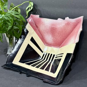 May include: A square ceramic bowl with a geometric design featuring a deep red interior, a cream-colored border, and a black base with parallel white stripes. The bowl has an irregular, textured rim and a glossy finish, showcasing a bold contrast.
