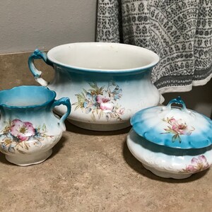May include: A set of antique porcelain bathroom fixtures. The set includes a large chamber pot, a small pitcher, and a lidded container. Each piece is white with a blue ombre rim and floral accents in pink, blue, and green.