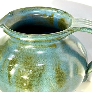 May include: A ceramic pitcher featuring a rounded body and a curved handle. The surface is finished with a glossy glaze in shades of light blue and earthy olive green, creating a mottled, blended appearance across the exterior and the rim.