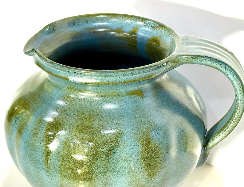 May include: A ceramic pitcher featuring a rounded body and a curved handle. The surface is finished with a glossy glaze in shades of light blue and earthy olive green, creating a mottled, blended appearance across the exterior and the rim.