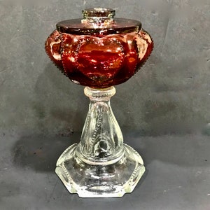 May include: A vintage glass oil lamp with a deep red, heart-shaped patterned reservoir and a clear glass base. The lamp has a decorative lid and a hexagonal base, showcasing a classic design.