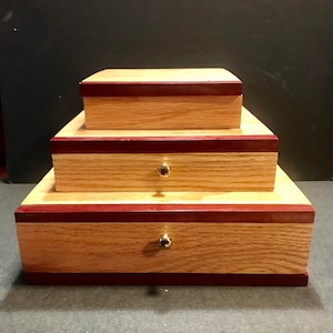 May include: A three-tiered wooden storage chest featuring light-colored oak wood panels accented with dark reddish-purple wood trim. The two lower levels function as drawers, each equipped with a small, round, polished gold-tone metal knob.