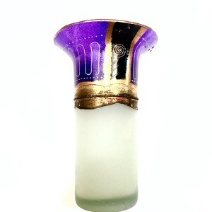 May include: A decorative glass bottle with a frosted body and a flared top. The top is decorated with purple, black, and gold designs. The bottle has a gold-colored metal band near the top.