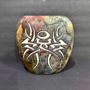 May include: A decorative, oval-shaped vase with a textured surface. The vase features a tribal design in white against a backdrop of earthy tones, including red, blue, and yellow. The design is centered on the front of the vase.