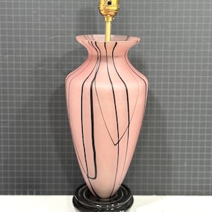 May include: A table lamp base featuring a matte pink glass body with vertical black line patterns. The lamp has a gold-colored metal socket at the top and sits on a circular, tiered black base. The design is tapered with a flared opening.