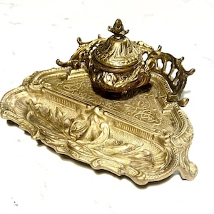May include: Ornate antique brass inkwell set. The set includes a detailed inkwell with a lid and a decorative tray. The inkwell and tray feature intricate embossed designs and a warm, golden hue. The tray has a rectangular shape with curved edges.