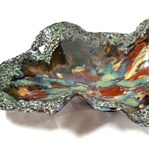 May include: A handcrafted ceramic serving dish with a unique, organic shape. The dish features a textured, dark green rim and a colorful interior with shades of red, blue, yellow, and brown. The dish is approximately 12 inches long.
