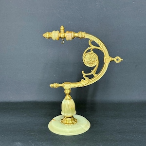 May include: A gold-toned metal display stand featuring an arched arm with ornate scrollwork and a small hanging hook. The base and central support are crafted from smooth, pale green stone, complemented by two small stone accents on the top arm.