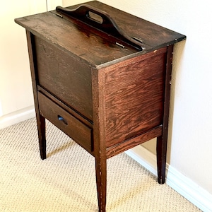 May include: A dark brown wooden side table with a drawer and a handle on the top. The table has four slender legs and is made of wood with a visible grain. The drawer has a small, round handle.