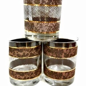 May include: Three clear drinking glasses with a brown leopard print design and gold trim. The glasses have a mesh pattern and are stacked in a triangular formation against a white background. The glasses are likely vintage.