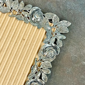 May include: A silver-colored metal picture frame with a floral rose and leaf design. The frame has a rectangular opening and is set against a brown cardboard background.