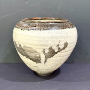 May include: A round ceramic vase with a wide opening and a tapered base. The exterior features a textured off-white glaze with dark brown, drip-like patterns running down the sides and a dark, mottled brown rim with a rough, weathered finish.