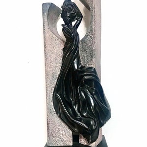 May include: A black sculpture of a figure with flowing fabric standing against a textured gray stone-like backdrop. The figure is mounted on a rectangular black base with visible wear along the bottom edge, creating a vertical composition.