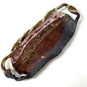 May include: An elongated ceramic tray with two twisted handles. The surface features a deep brown and black glaze with a textured, raised band of white and pinkish-purple detailing running along one side, accented by small, dark, circular indentations.