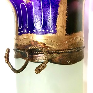 May include: A decorative glass vase with a frosted white body and a detailed top. The top features a vibrant blue and gold design with white accents. A bronze-colored rope detail adds a rustic touch to the vase.