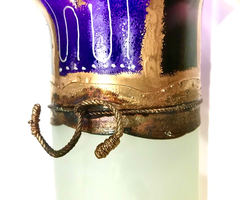 May include: A decorative glass vase with a frosted white body and a detailed top. The top features a vibrant blue and gold design with white accents. A bronze-colored rope detail adds a rustic touch to the vase.