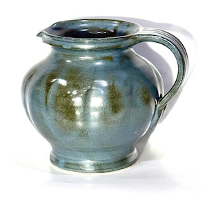 May include: A handmade ceramic pitcher with a blue and green glaze. The pitcher has a rounded body, a spout, and a curved handle. The glaze has a mottled appearance, with variations in color. The pitcher is suitable for serving beverages.