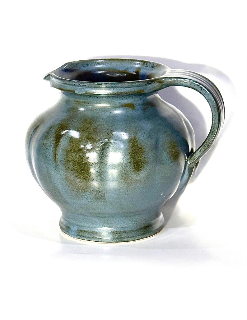 May include: A ceramic pitcher with a rounded body and a wide, flared rim. The surface features a glossy glaze in shades of blue and olive green, with vertical indentations along the sides and a curved handle attached to the side of the vessel.