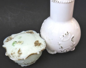 May include: A white milk glass vase with a spherical body and embossed scrollwork details, paired with a matching lidded jar featuring gold-painted accents, scalloped edges, and floral motifs on the lid surface.