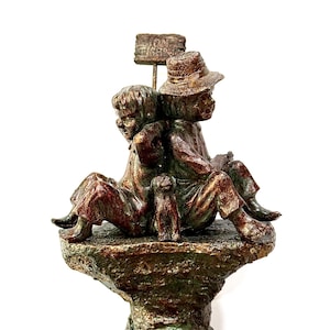 May include: A bronze-toned sculpture featuring two figures sitting back-to-back on a textured rock base with a small dog between them. A sign above the figures reads GONE FISHING. The sculpture has a weathered finish with green and brown highlights.