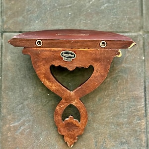 Puede incluir: Una estanter&iacute;a de madera vintage con un dise&ntilde;o recortado en forma de coraz&oacute;n. La estanter&iacute;a es marr&oacute;n con detalles dorados y presenta la etiqueta "Syroco Wood". Tiene dos ojales color lat&oacute;n para colgar.