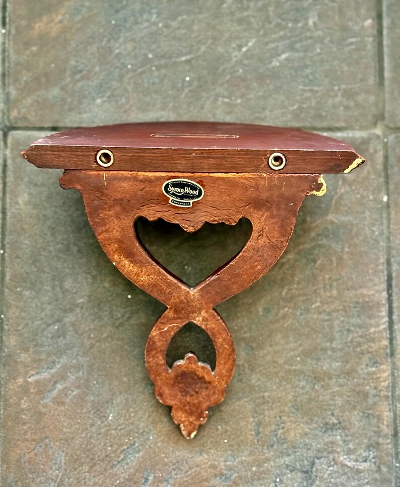 Puede incluir: Una estanter&iacute;a de madera vintage con un dise&ntilde;o recortado en forma de coraz&oacute;n. La estanter&iacute;a es marr&oacute;n con detalles dorados y presenta la etiqueta "Syroco Wood". Tiene dos ojales color lat&oacute;n para colgar.