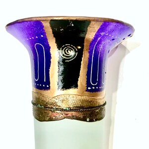 May include: A decorative vase with a flared top. The top is painted with purple, black, and gold sections, accented with white lines and a spiral design. The base is a frosted white color.