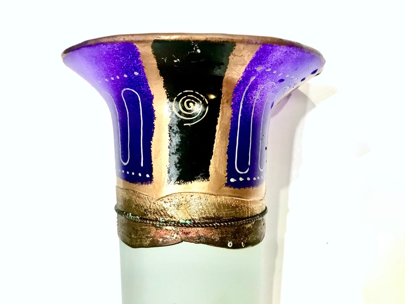May include: A decorative vase with a flared top. The top is painted with purple, black, and gold sections, accented with white lines and a spiral design. The base is a frosted white color.