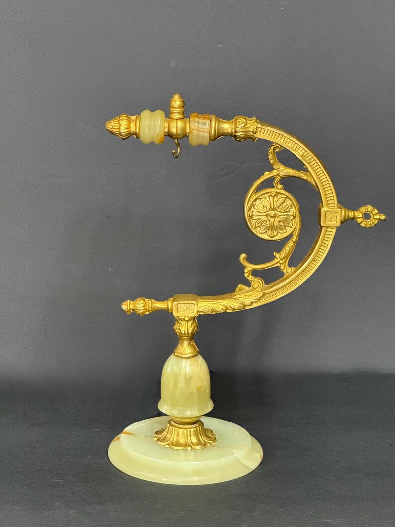 May include: A gold-toned metal stand featuring a curved arm with ornate scrollwork and a small hanging hook. The base and central support are crafted from pale green onyx stone, accented with gold-toned metal detailing and decorative finials.