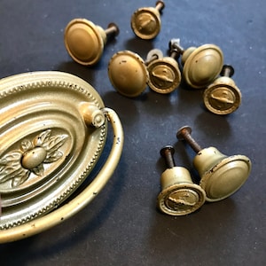 May include: A collection of antique brass cabinet hardware, including several round knobs and an oval pull with a floral design. The hardware has a distressed, aged appearance with visible wear and tear, suggesting vintage origins.