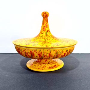 May include: A yellow ceramic lidded dish with a pedestal base. The dish and lid are decorated with a speckled pattern of red, green, and yellow. The lid has a tall, conical handle.