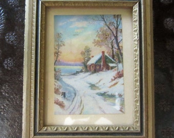 May include: A rectangular framed print featuring a winter landscape with a small cabin, snow-covered ground, bare trees, and a sunset sky. The frame is gold-toned with a textured border and a small white pull tab at the bottom center of the frame.