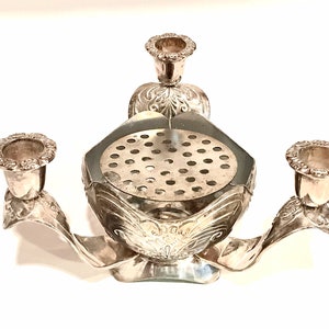 May include: A silver-toned metal centerpiece featuring three candle holders with scalloped edges and a central bowl with a perforated insert. The base and candle holders are decorated with embossed floral and leaf-like patterns throughout.