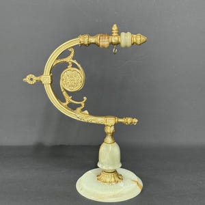 May include: A gold-toned metal display stand featuring an ornate curved arm with decorative scrollwork and a circular medallion. The base and accents are crafted from pale green and white banded stone, including a small hanging hook.