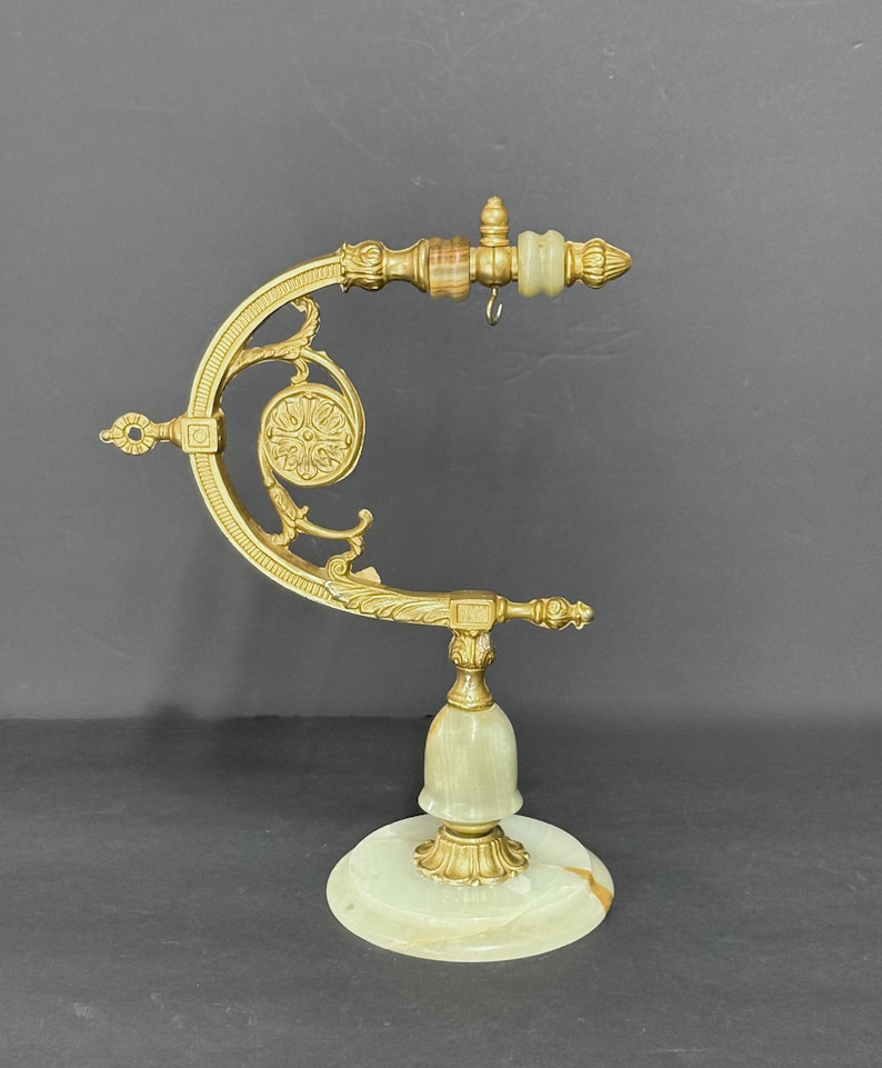 May include: A gold-toned metal display stand featuring an ornate curved arm with decorative scrollwork and a circular medallion. The base and accents are crafted from pale green and white banded stone, including a small hanging hook.