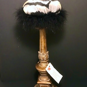 May include: A round pincushion with a black and white abstract pattern, trimmed with black feathers, resting on a tall, weathered bronze-colored pedestal stand. A single decorative pin with a white bead is inserted into the top of the cushion.