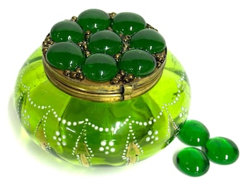 May include: A round, lime green glass trinket box with a brass lid featuring seven embedded dark green glass cabochons. The glass body is decorated with white painted dots and gold arrow-like motifs. Three loose dark green glass stones sit nearby.