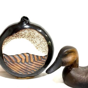 May include: A circular ceramic vase with a glossy black outer rim, a speckled white center, and a textured brown base with horizontal ridges, positioned next to a carved wooden duck figurine with a dark brown finish and a black beak.