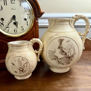 May include: A cream-colored ceramic pitcher and a smaller matching creamer, both featuring gold-toned trim and a brown-toned illustration of a person in profile wearing a head covering. Each piece has a decorative handle shaped like a dragon head.